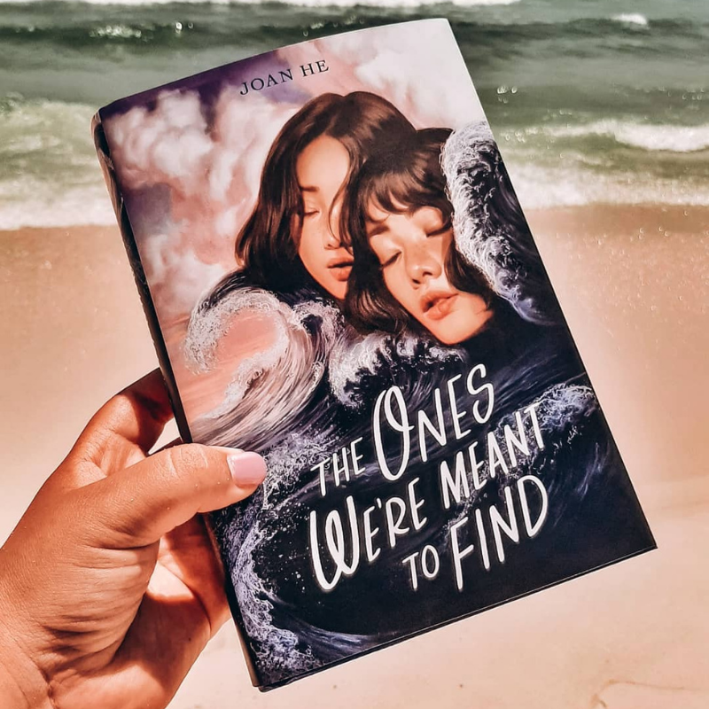 A hand holding a copy of The Ones We're Meant to Find on a sandy beach in front of the ocean. 