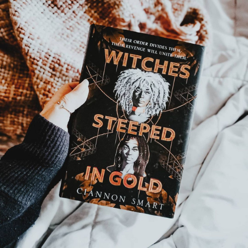 A hand holding a copy of Witches Steeped in Gold over a white blanket and a knitted throw blanket