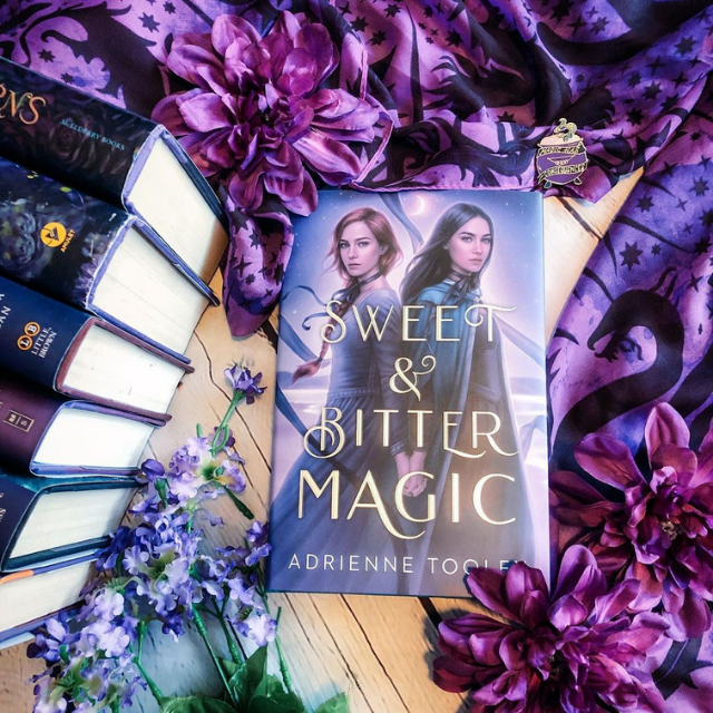 A copy of Sweet & Bitter Magic laying flat surrounded by purple flowers, a purple scarf, and purple book spines.