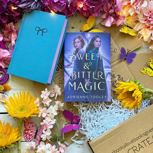 Two copies of Sweet & Bitter Magic, one without a dust jacket, laying flat on an open cardboard OwlCrate box, surrounded by colourful flowers