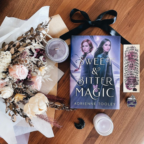 A copy of Sweet & Bitter Magic laying flat beside a bouquet of flowers on a wooden surface along with two candles and a black ribbon tied in a bow.