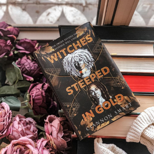 A copy of Witches Steeped in gold laying on top of book spines beside some pink flowers