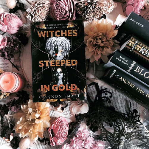 A coping of Witches Steeped in Gold surrounded by mostly pink flowers and book spines