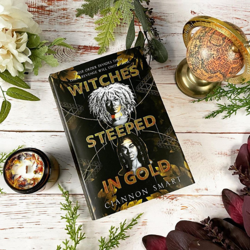 A copy of Witches Steeped in gold laying on white wooden boards surrounded by a globe, a candle, and some flowers