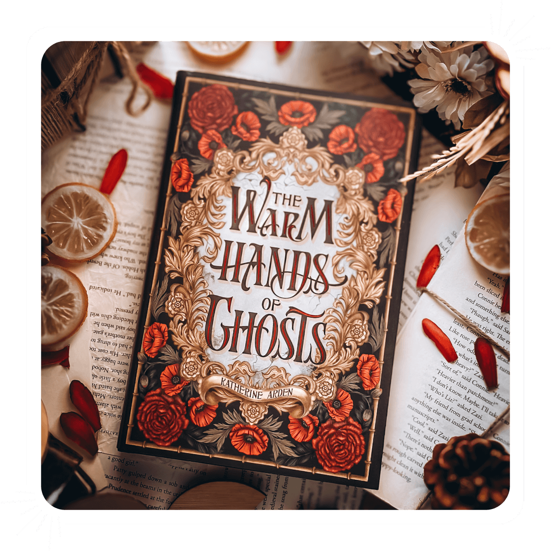 OwlCrate exclusive edition of The Warm Hands of Ghosts by Katherine Arden lying on a table, surrounded by book pages, red petals, and orange slices. The book cover features roses, poppy flowers, and a golden frame around the book title.