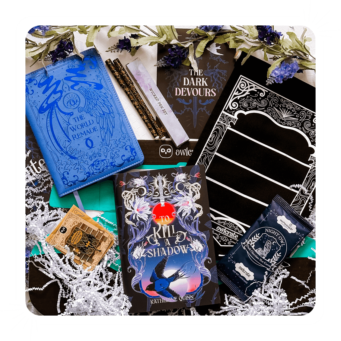 Items from an OwlCrate Young Adult subscription box lying on a table with purple flowers above them. Items featured include a book-shaped pin, a blue journal with text reading The World Remade, two black and gold pens with a box next to them, a theme reveal card reading The Dark Devour, a black pin banner with a white design around it, and OwlCrate written at the bottom, a pack of hot chocolate that says Night Owl, and the To Kill A Shadow book in the bottom middle.