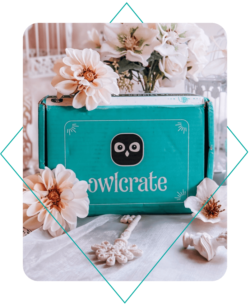 A teal box with a white border, an OwlCrate logo in the center, and white text under it reading OwlCrate. The box is on white fabric and is surrounded by white flowers.