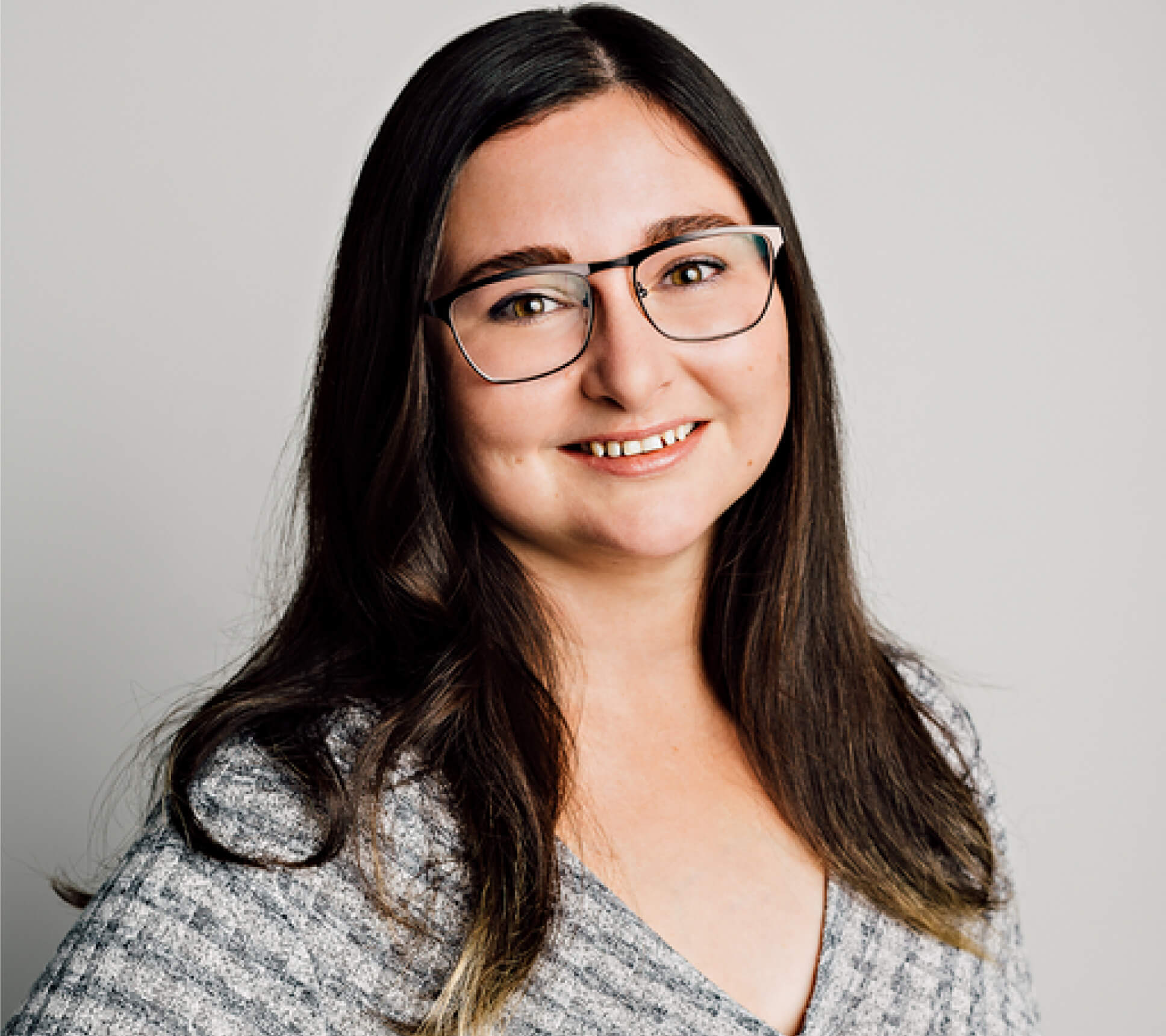 A white woman with brown eyes and long brown hair, smiling with her head slightly tilted to the side, wearing metal black rectangular glasses and a grey shirt with a dark grey and light grey lattice design.